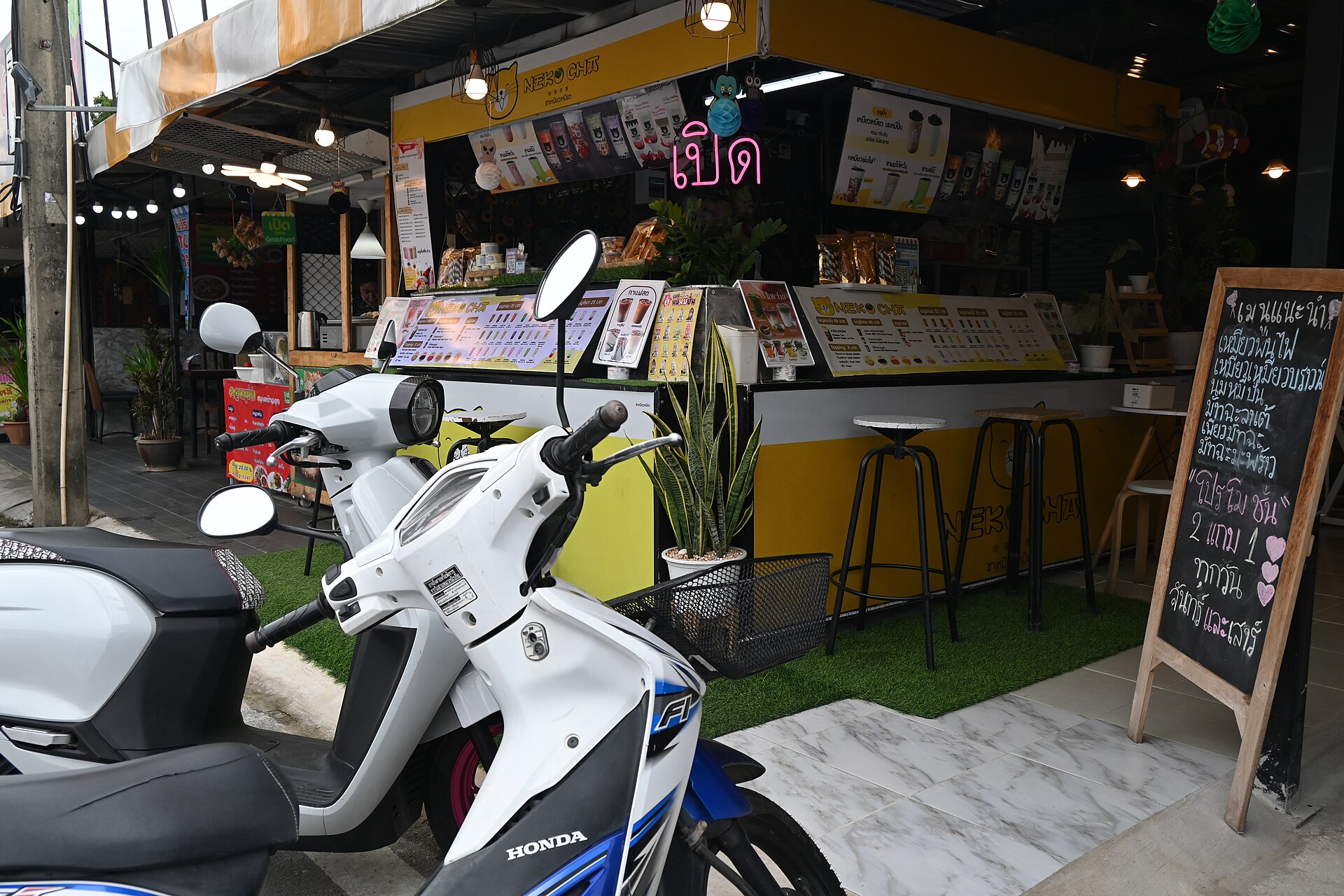 DFC 2419 A bright white and blue Honda scooter parked in front of a cozy yellow street side drink shop with menu boards and stools
