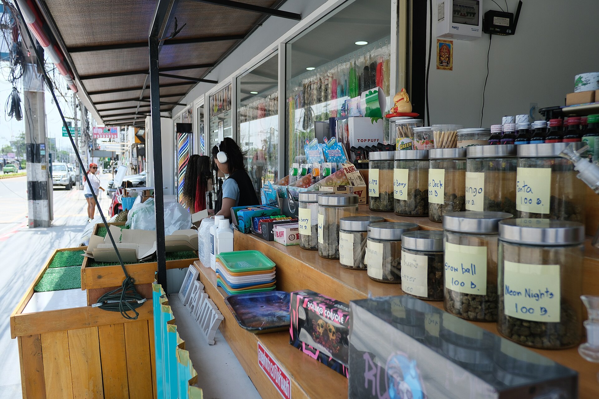 DSCF0824 A colorful street side shop display with jars of snacks and small goods lined up on a wooden counter while a sales attendant chats with customers under a shaded awning