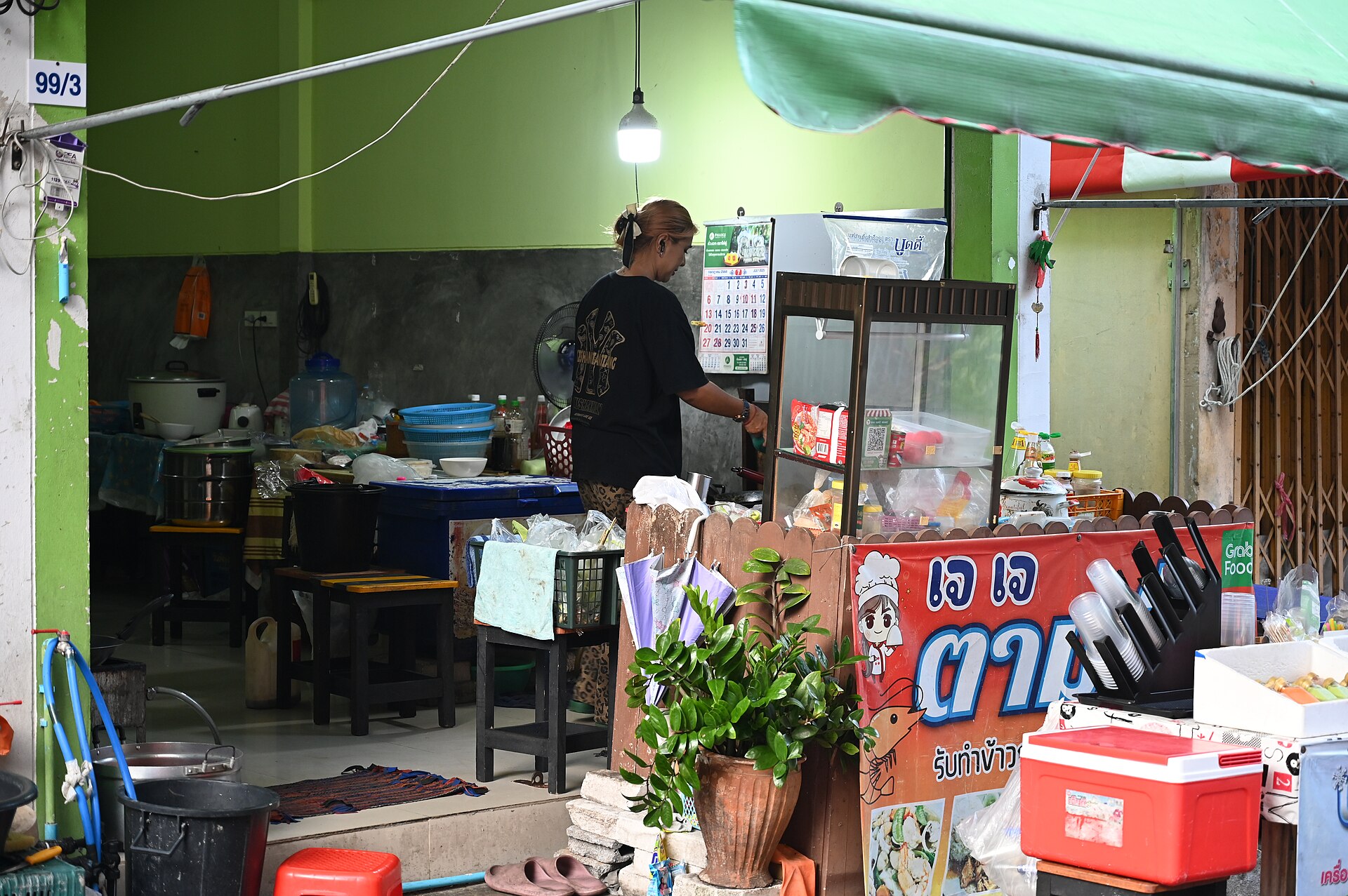 DZ6 0527 A small street side eatery with a vendor preparing food behind a glass display case surrounded by pots bowls and potted plants