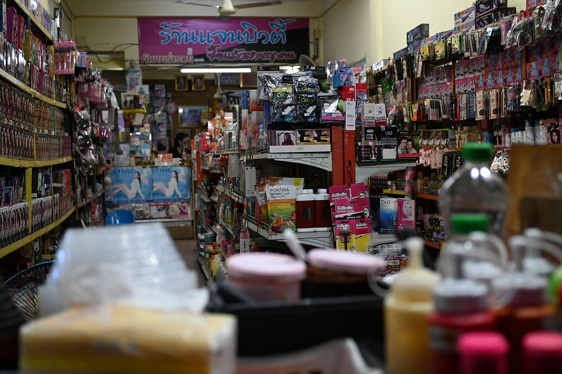 DFC 3031 A narrow bustling shop aisle packed from floor to ceiling with colorful household goods toiletries and snacks stacked on shelves under a sign in Thai