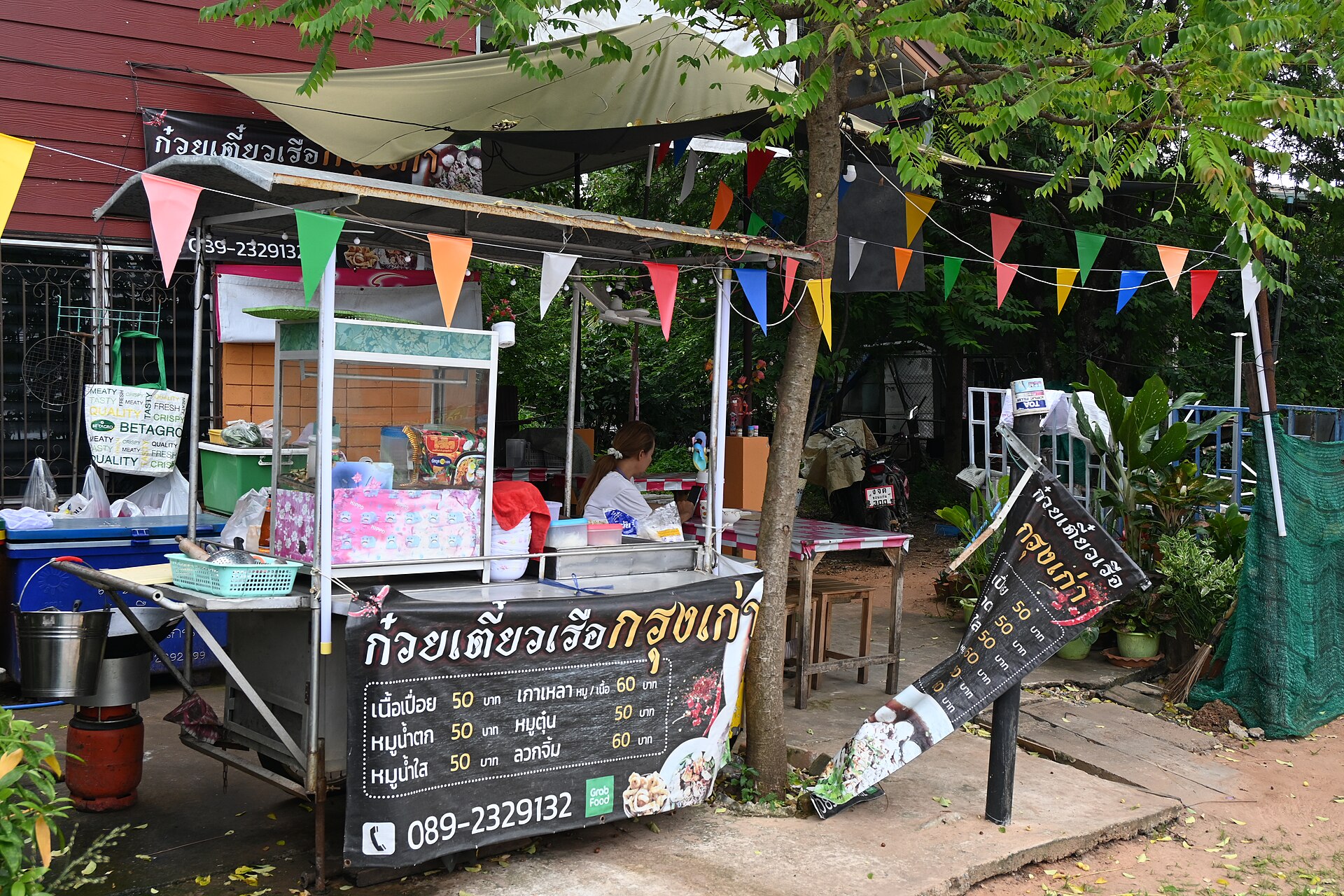 DFC 2418 A small outdoor street food stall with colorful pennant flags a display case of ingredients and a handwritten menu sign in Thai beside the sidewalk