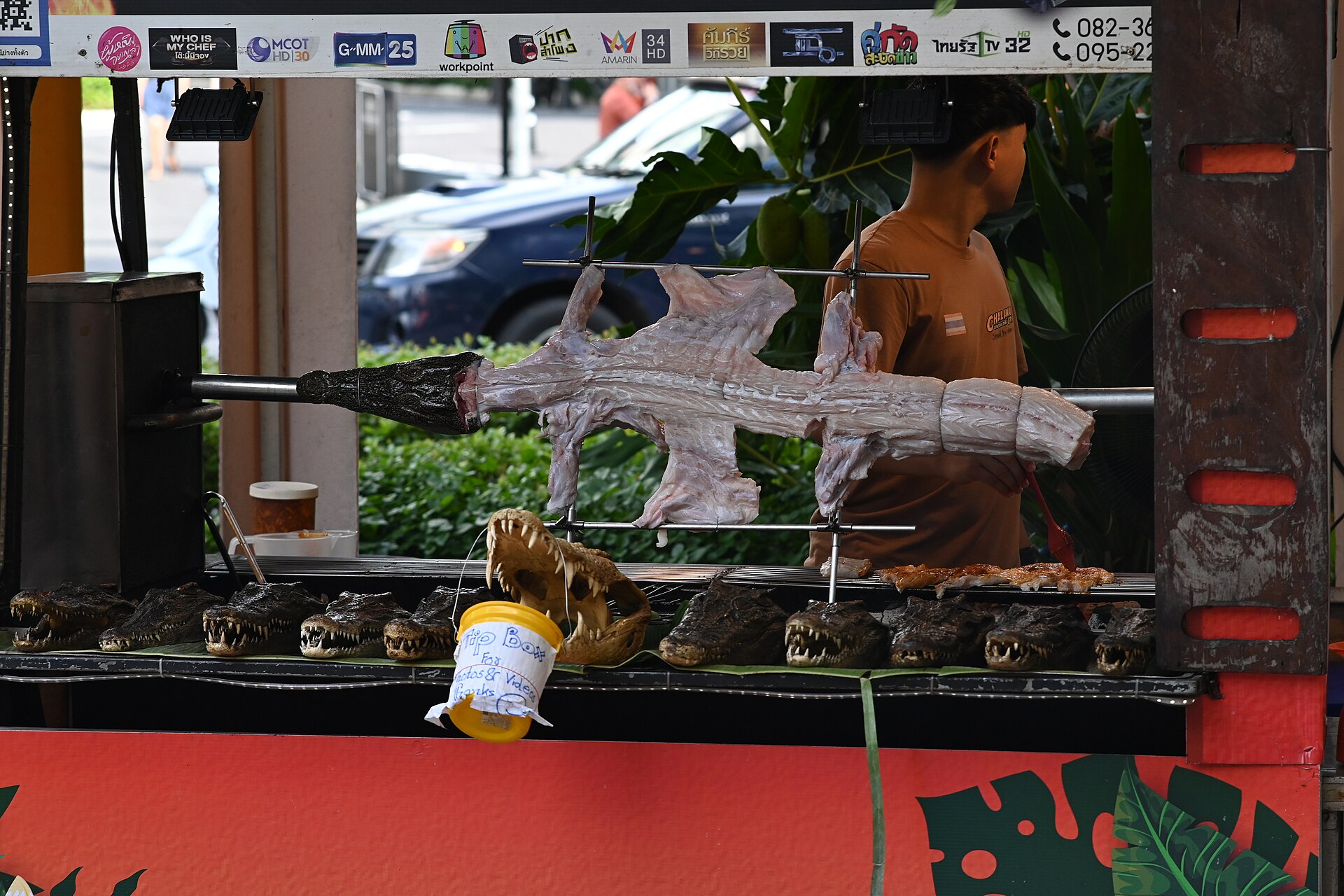 DFC 0838 A whole crocodile roasting on a spit at an outdoor food stall with small crocodile heads arranged on the grill and a vendor standing behind the counter