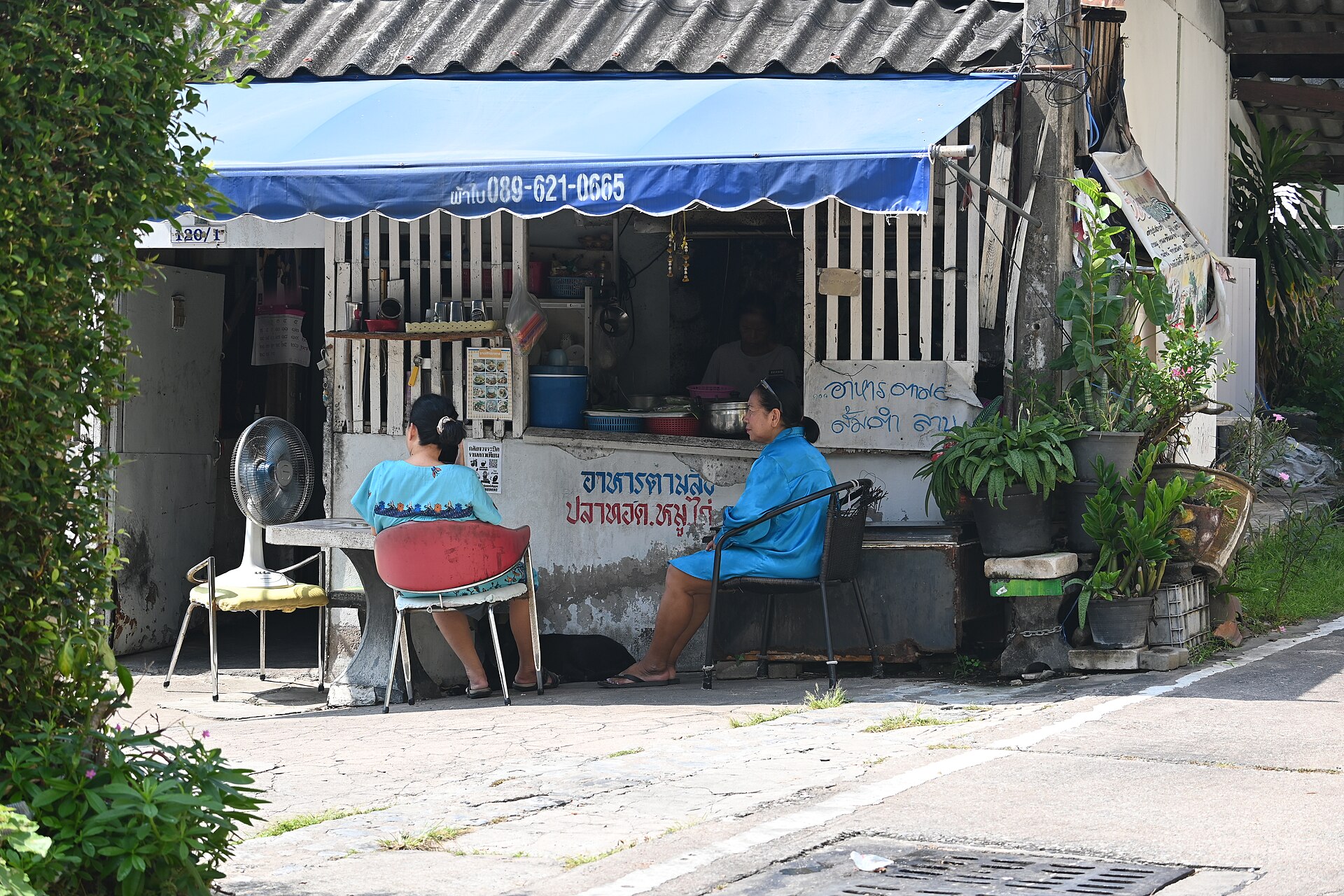 DFC 2053 Two friends chat over drinks outside a small neighborhood stall shaded by a blue awning
