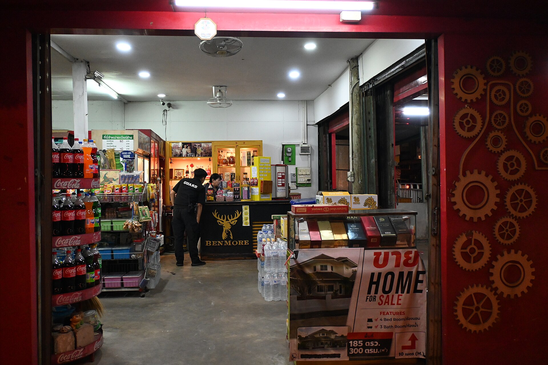 DFC 2489 A brightly lit convenience store at night with shelves of bottled drinks and snacks a stack of water bottles and a Home for Sale sign near the entrance and a person behind the counter