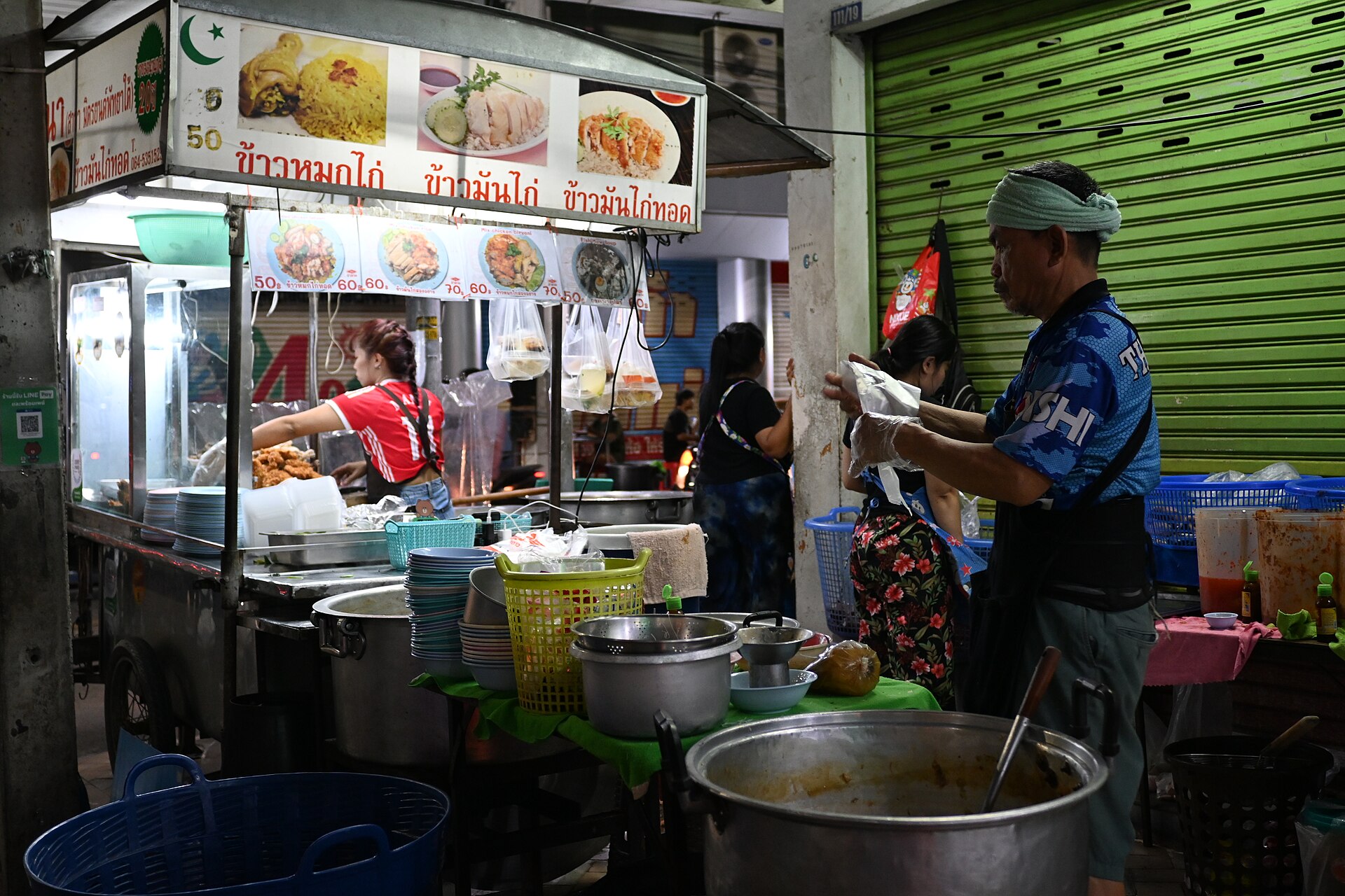DFC 1643 A busy Thai street food stall at night with a vendor preparing dishes behind the counter and a customer waiting as large pots and ingredients fill the scene