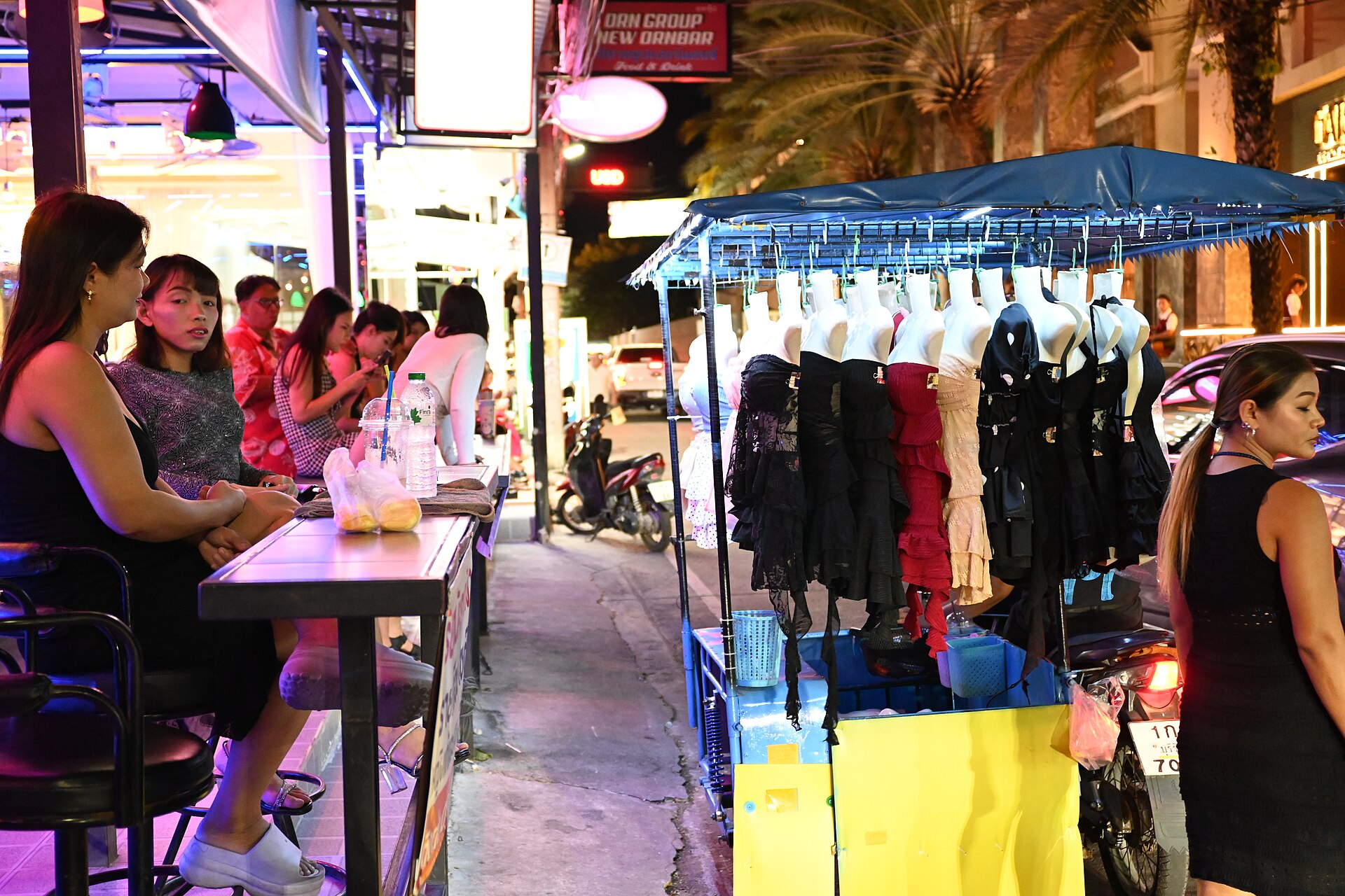 DFC 1681 Nighttime street scene with a small outdoor stall displaying dresses under a canopy while people sit and chat at a nearby bar lined sidewalk