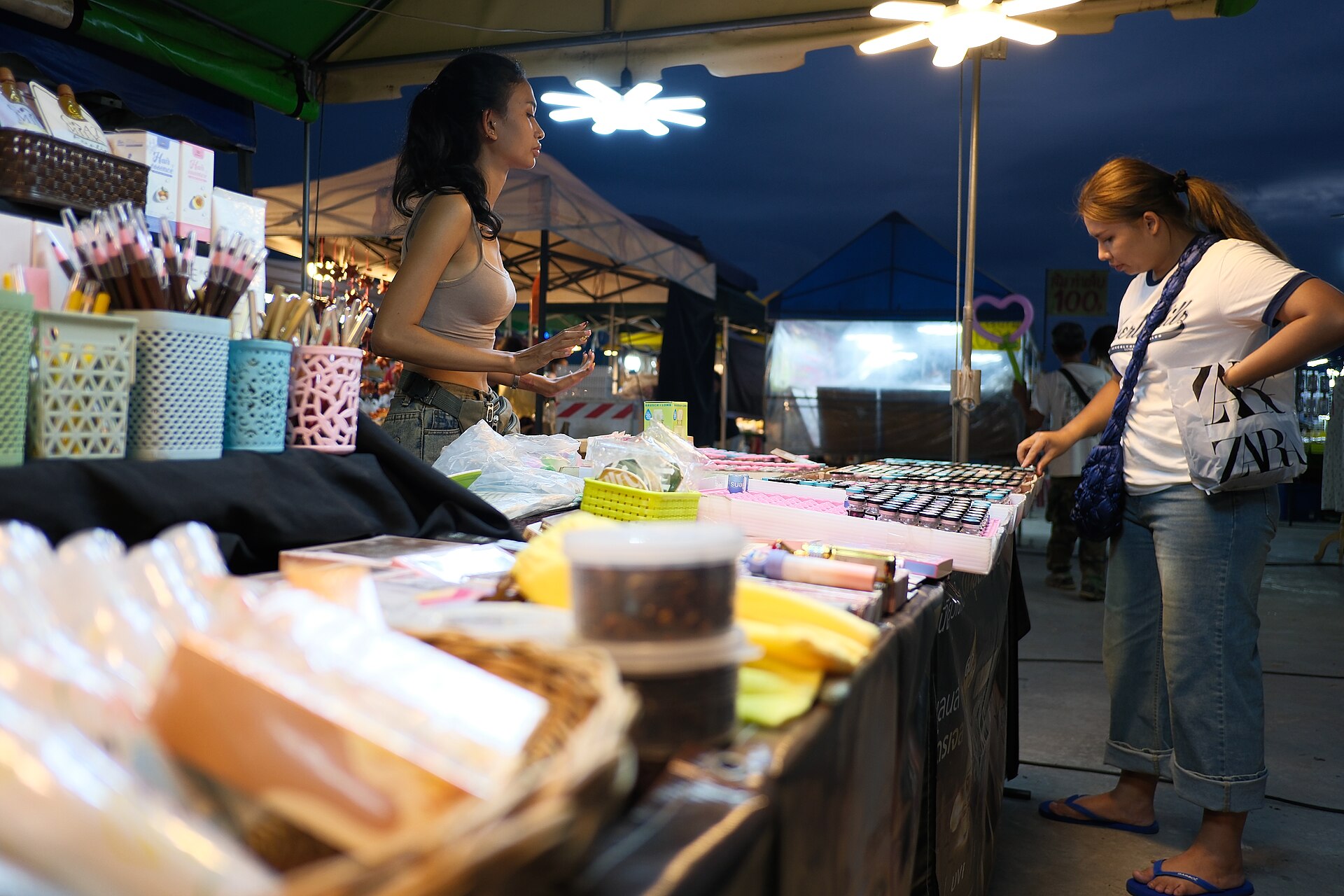 DSCF0909 Two women browse and chat at a brightly lit outdoor market stall displaying an array of small goods and cosmetics at dusk