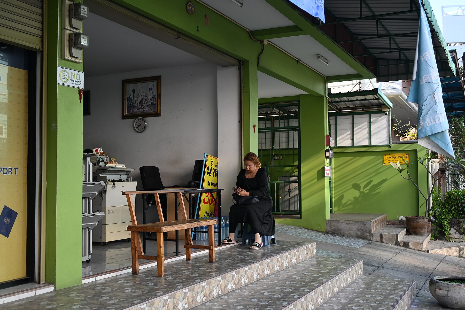 DFC 4008 A woman sits on the steps outside a small green storefront checking her phone beside a wooden chair and a modest office setup