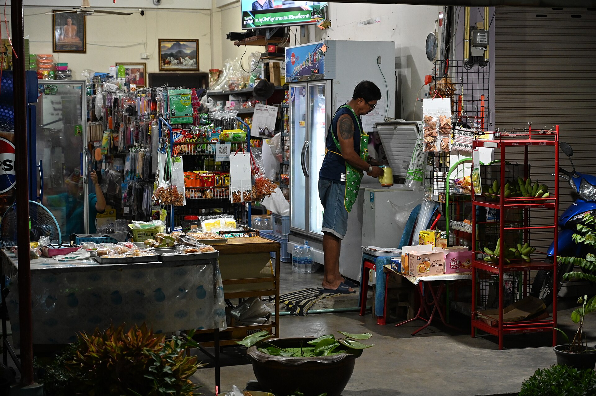 DZ6 0569 A small neighborhood shop at night with a vendor preparing items behind the counter shelves stocked with snacks and household goods and fresh produce on display
