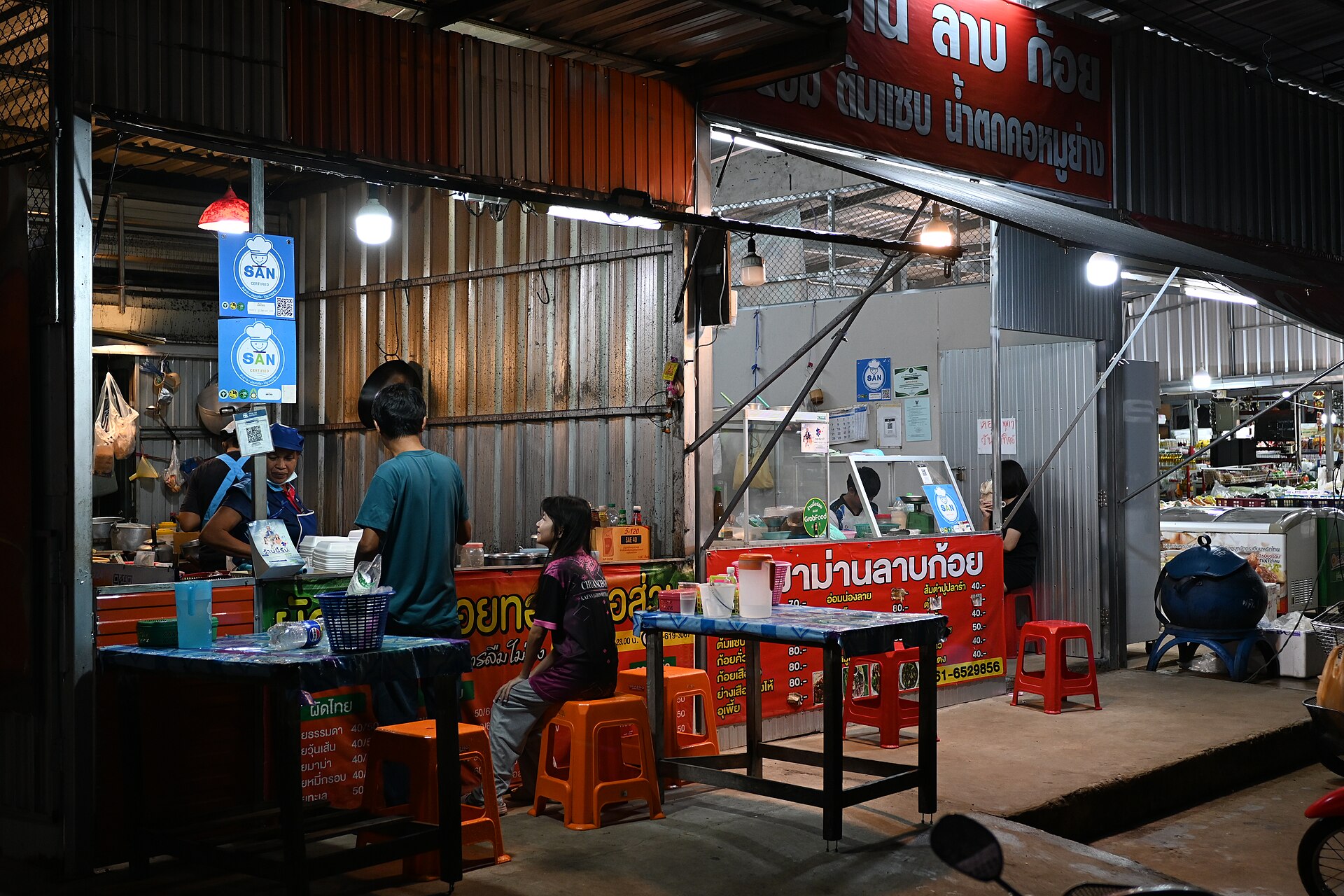 DFC 2557 A small street food stall lit warmly at night with customers seated on plastic stools as vendors prepare meals behind the counter