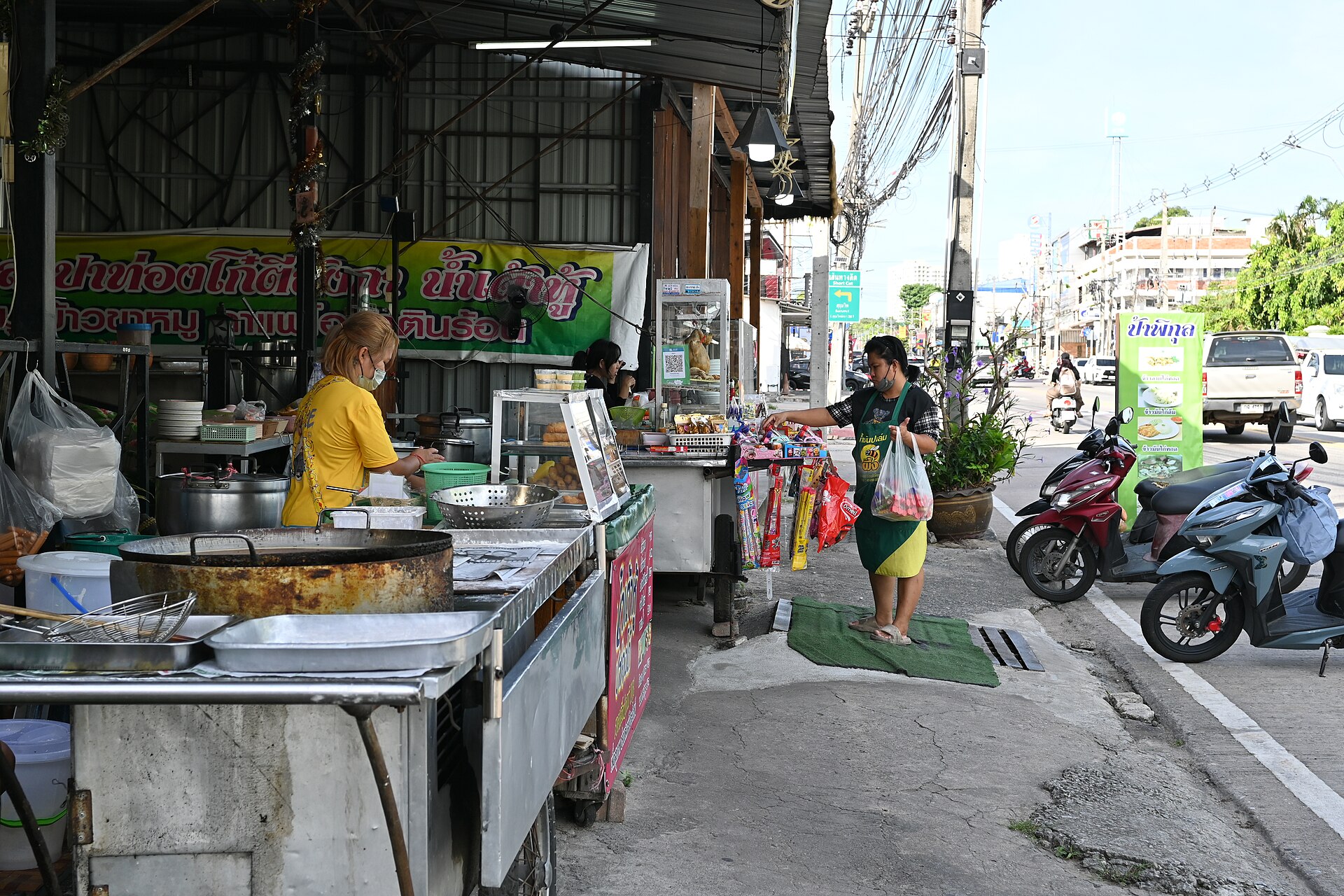 DFC 1271 A street food vendor in a yellow shirt prepares dishes at an open air stall while a customer carrying bags approaches along the sidewalk motorbikes are parked nearby and buildings line the busy street