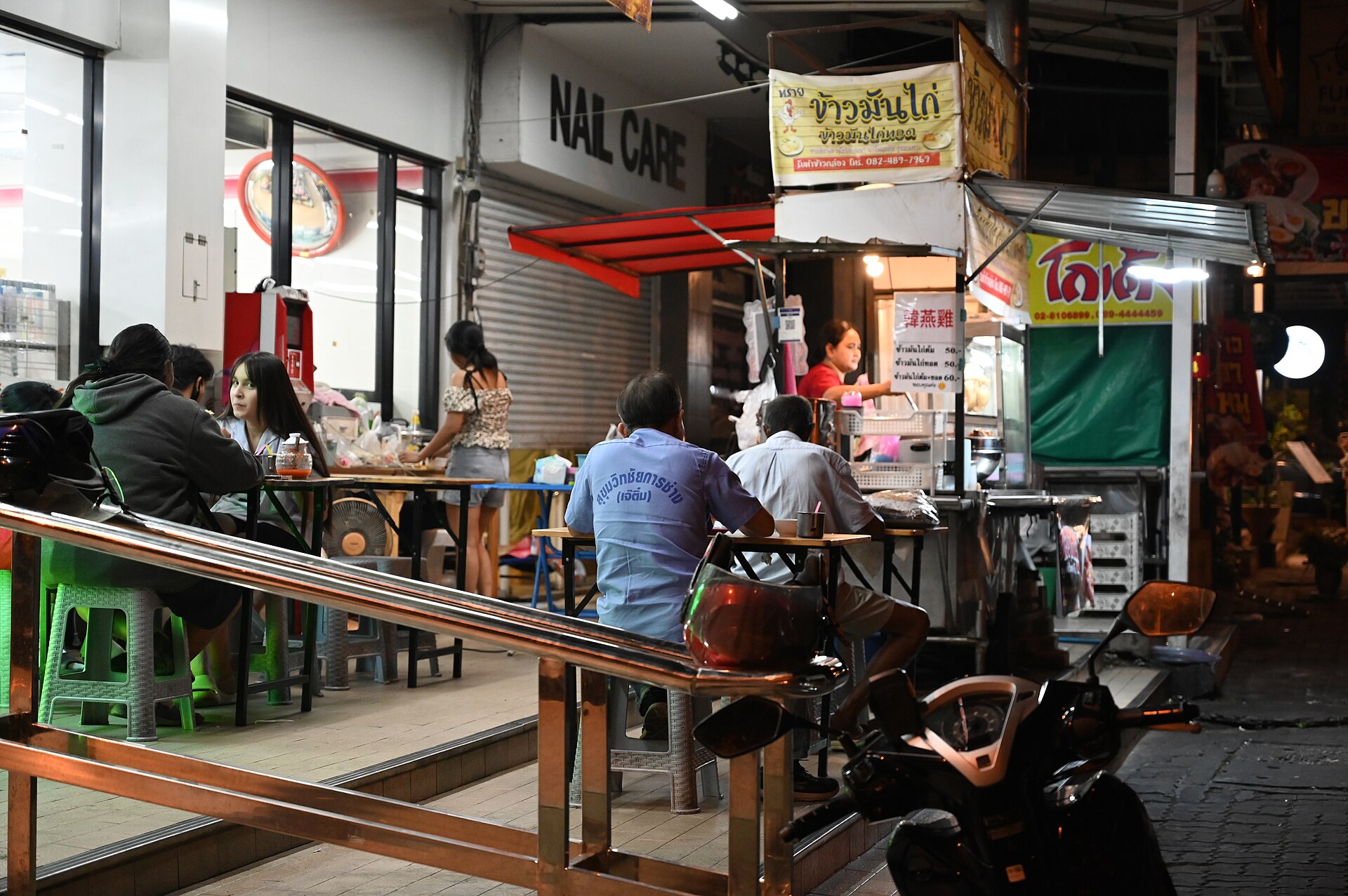 DSC 8540 A nighttime street food stall draws locals seated at plastic tables and stools outside a small shop with a vendor preparing food under bright lights and a parked motorbike in the foreground